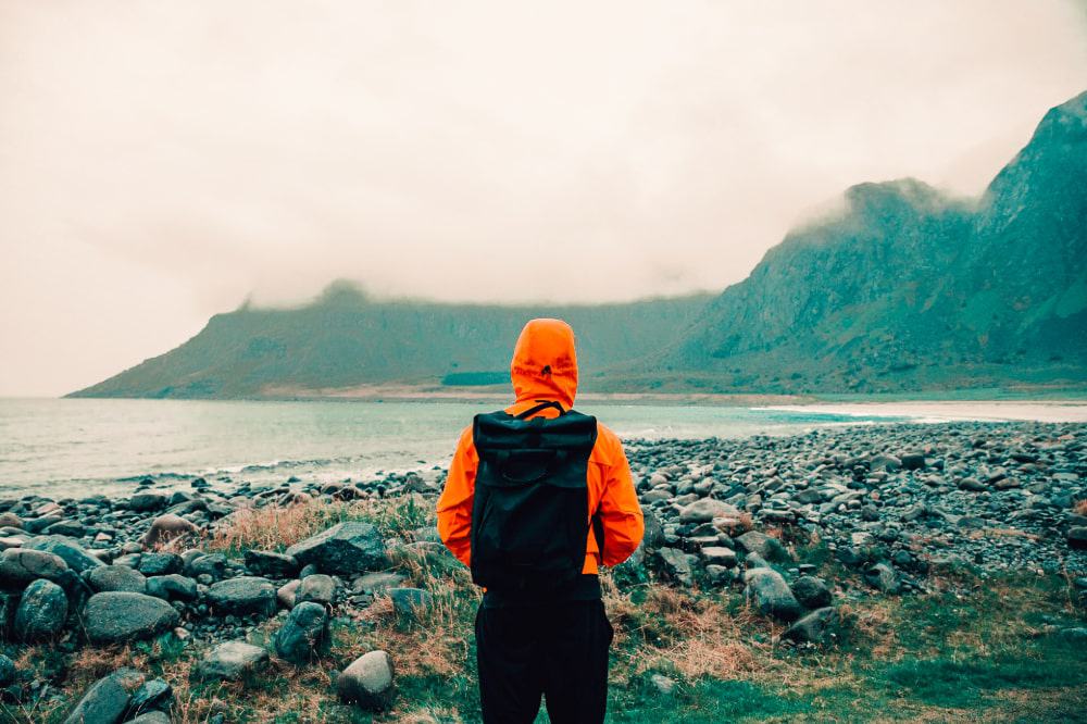 A person in an orange rain jacket looks at hills on a rocky shore