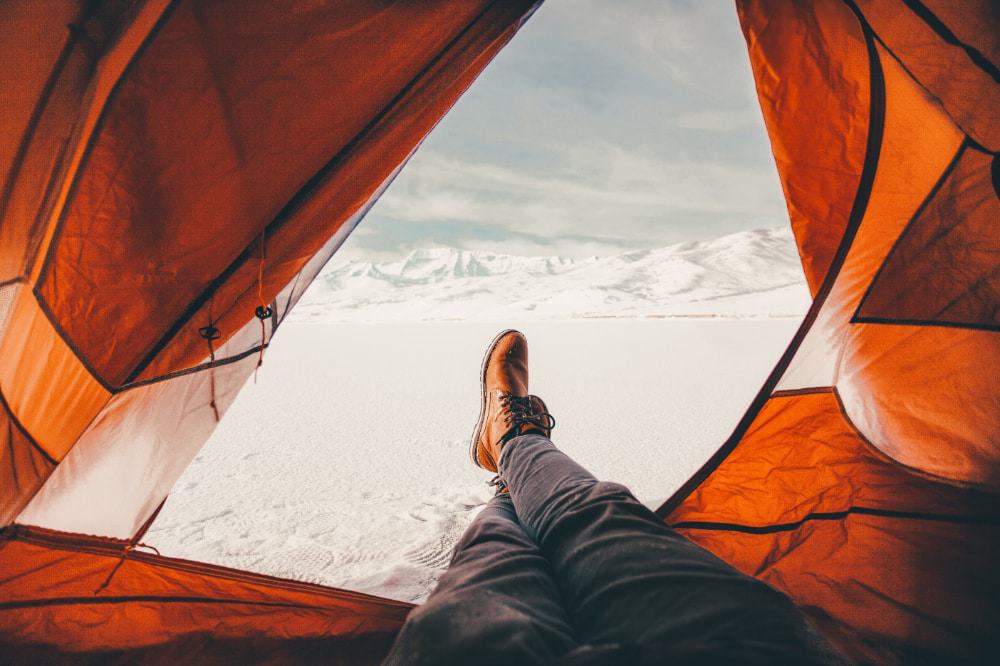 A person sat with their feet out of their open tent looking snow-covered mountains 